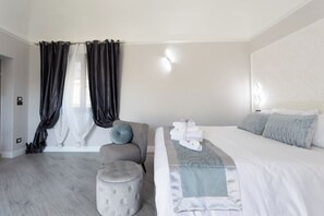 A modern bedroom with a large bed, a grey armchair, a small ottoman, and a window with black curtains. - Cozy studio apartment within walking distance of hospital (Vercelli)
