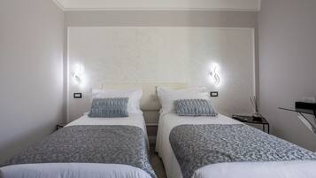A hotel room with two beds, each with a patterned bedspread, white pillows, and a headboard. There are wall-mounted lights on either side of the bed, and a small table with a vase and a remote control.