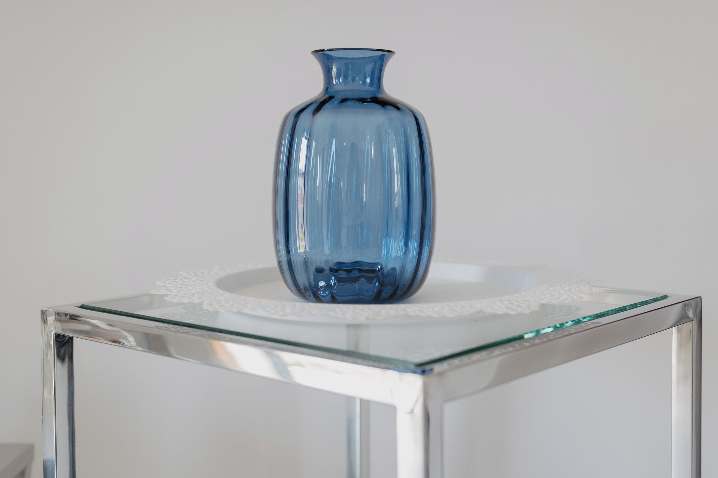 Un florero azul de vidrio sobre una mesa de vidrio transparente con un mantel de encaje.