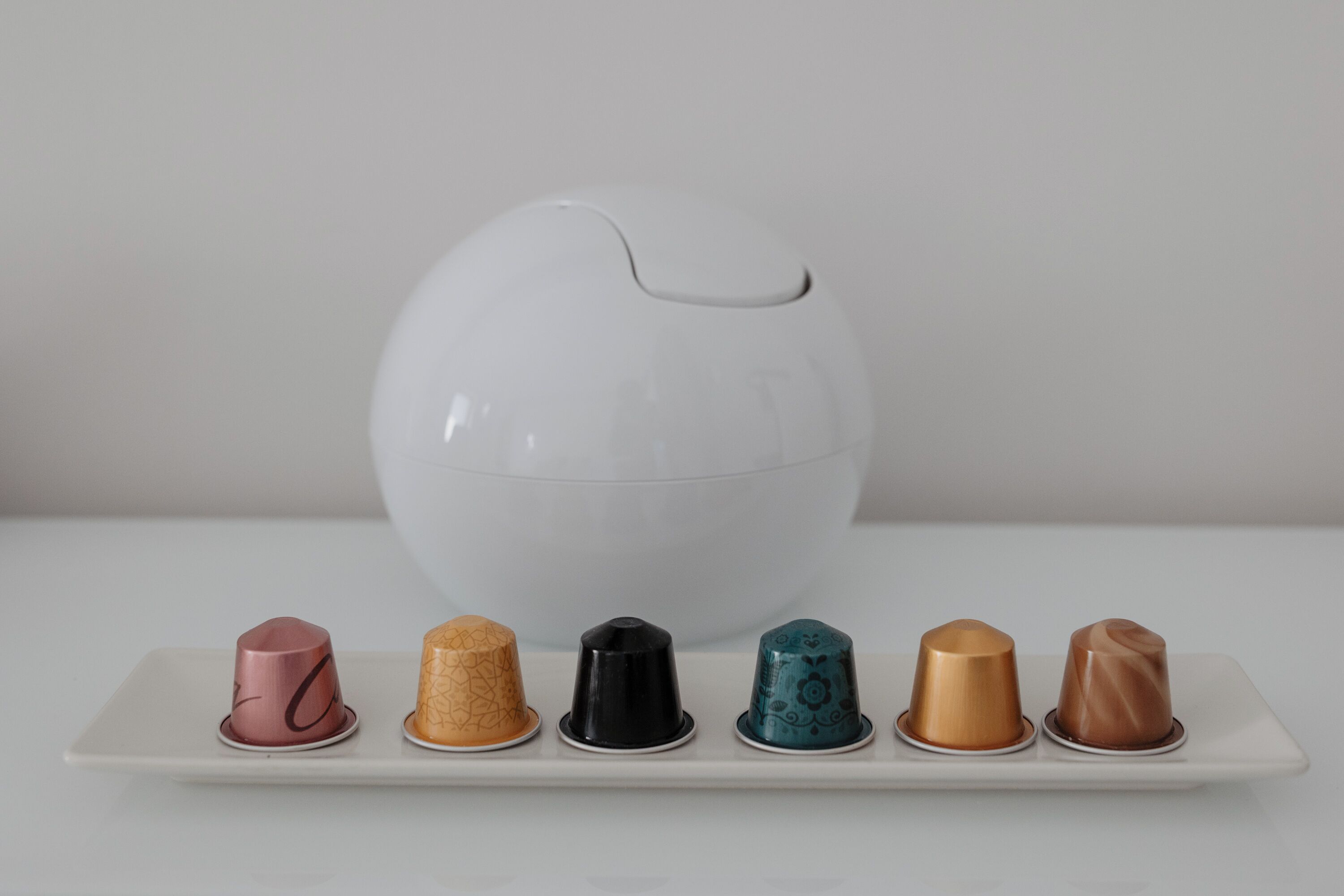 A white ceramic egg-shaped object on a white surface with a white plate below it containing six differently colored and patterned cups.