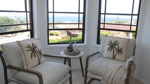 Deux chaises ornées de motifs de palmiers, une petite table sur laquelle est posé un objet décoratif, et une vue sur l’océan par de grandes fenêtres.