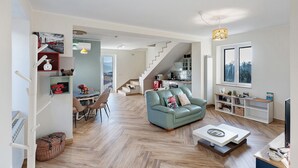Ein modernes Wohnzimmer mit einer Couch, einem Couchtisch und einem Fernseher. Eine Treppe führt zu einer anderen Ebene, im Hintergrund sind ein Essbereich und eine Küche zu sehen.