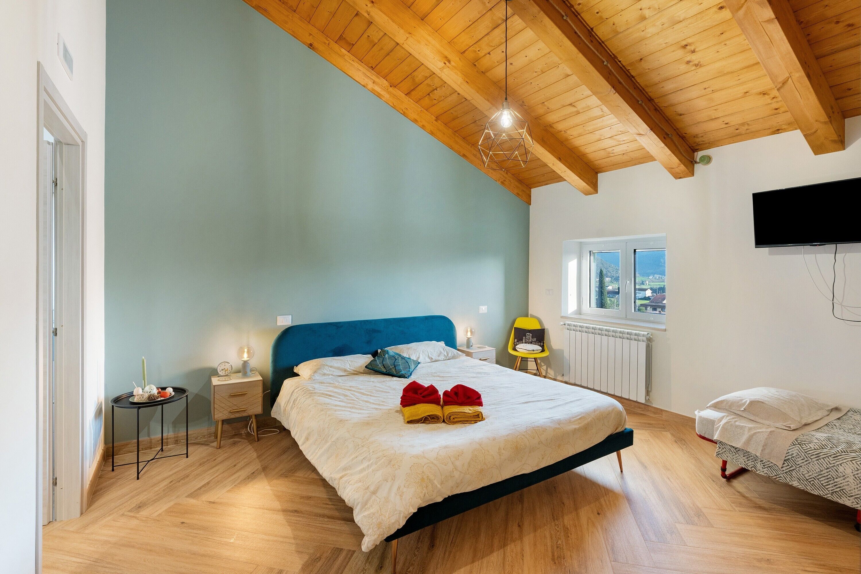 A bedroom with a wooden ceiling, a bed with a blue headboard, a bedside table with a lamp, and a window with a view.