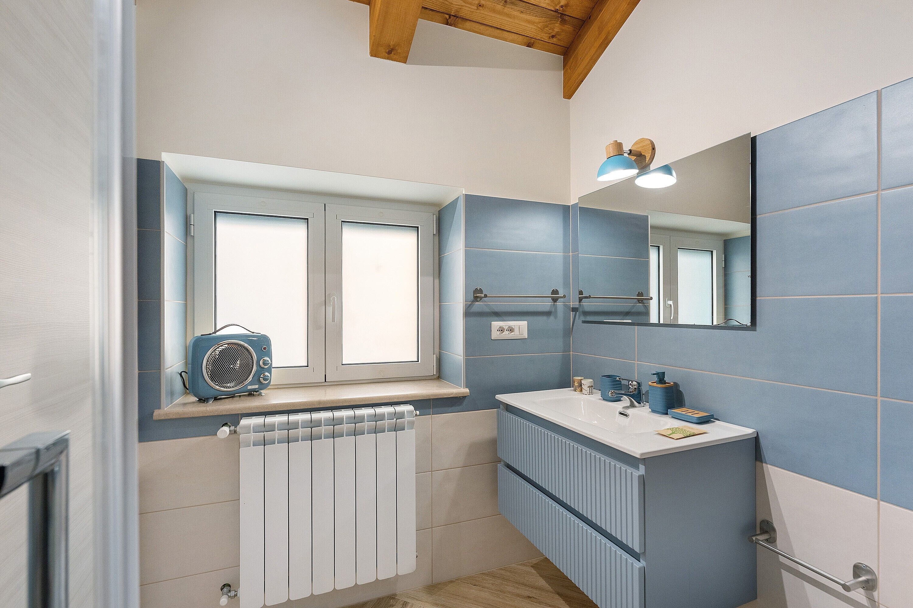 A bathroom with a window, a radiator, a sink, a mirror, and a blue-tiled wall.