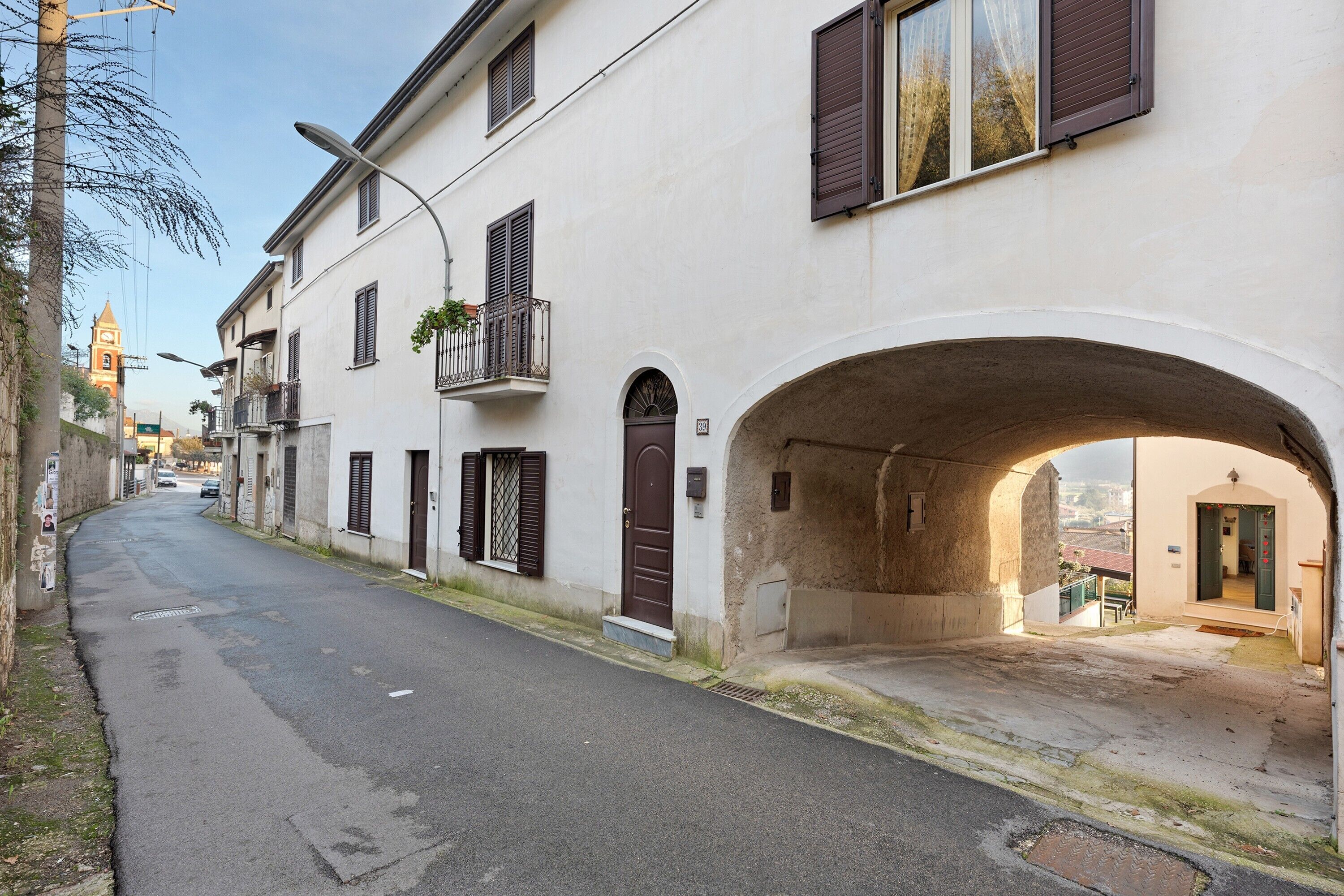 Una stretta via con un edificio bianco caratterizzato da un'ampia arcata, una porta marrone e un piccolo balcone con piante.