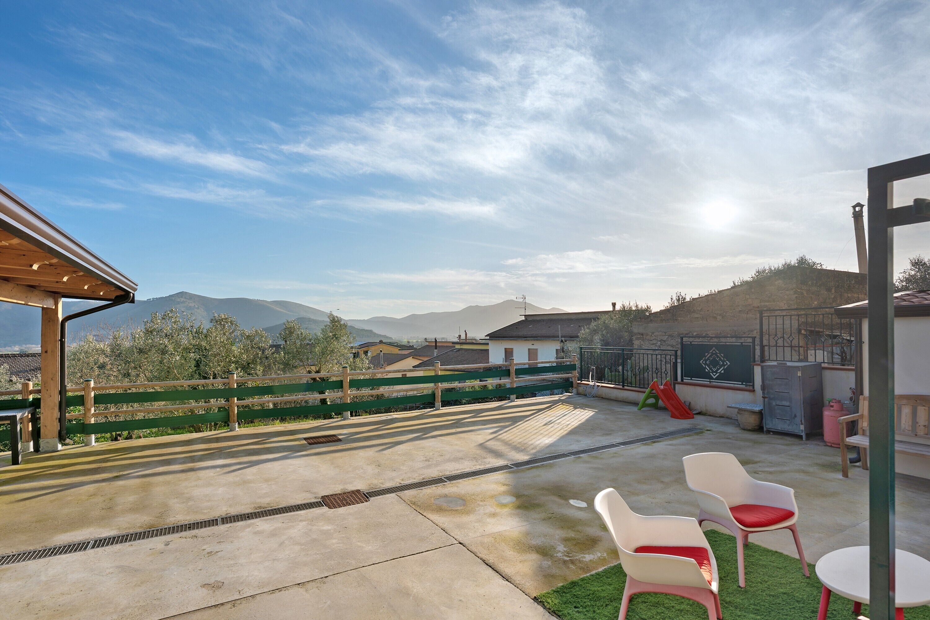 Un patio en la azotea con piso de concreto, dos sillas de plástico blanco con cojines rojos, una mesa blanca pequeña y un tobogán verde.