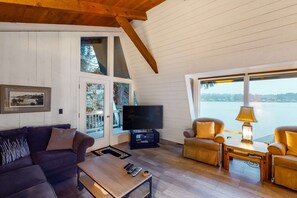 Una sala de estar acogedora con techo de madera, un ventanal grande con vistas al agua, un televisor y asientos cómodos.