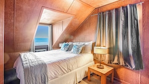 Un dormitorio con techo inclinado, una cama con edredón blanco, dos almohadas, una mesita de noche con lámpara y una ventana con cortinas.