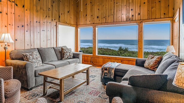 Una habitación acogedora con paredes de madera, amplias ventanas y vistas al océano, un sofá, una mesa de centro y una lámpara.
