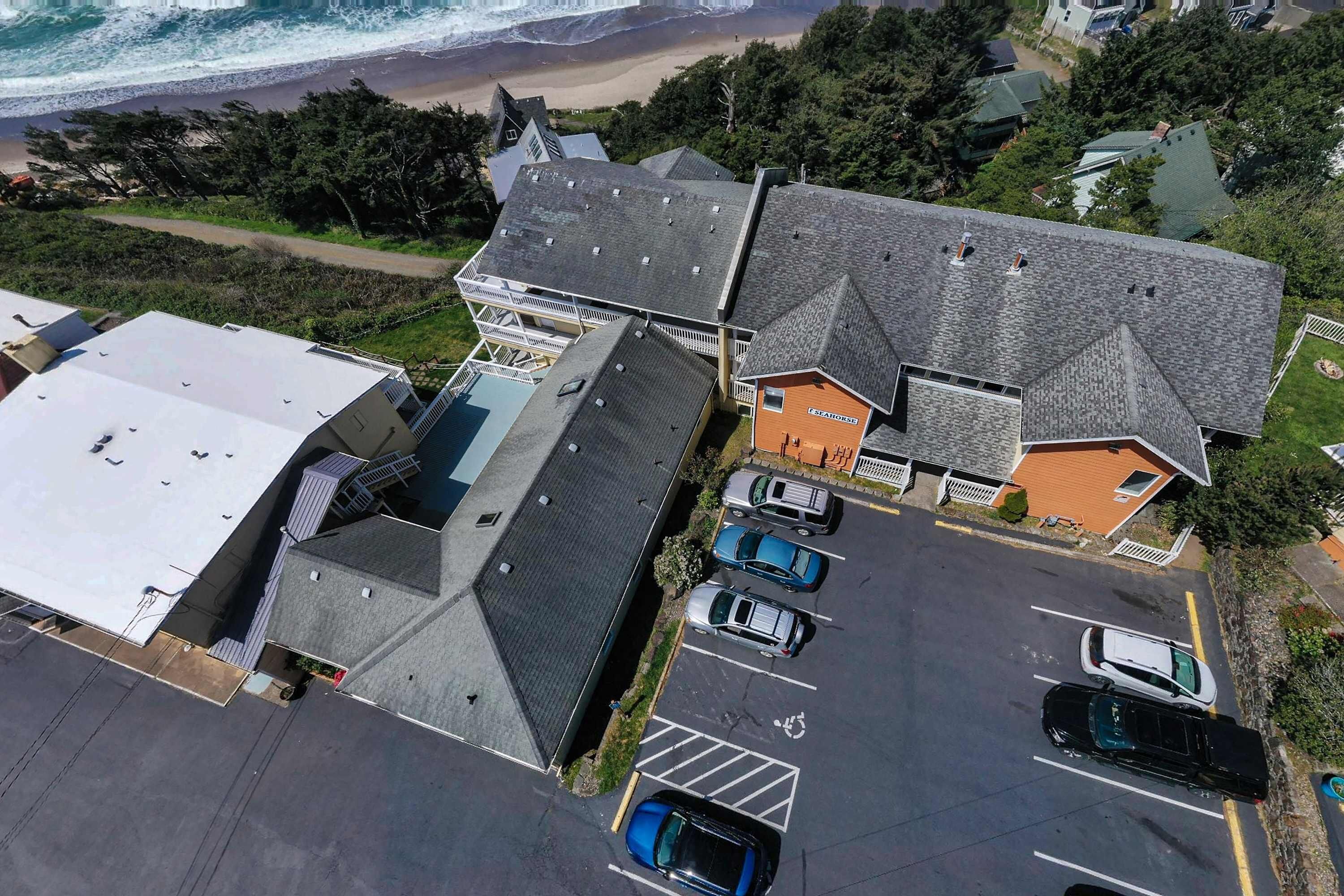 An aerial view of a coastal property with multiple buildings, a parking lot with several cars, and a beach in the background.
