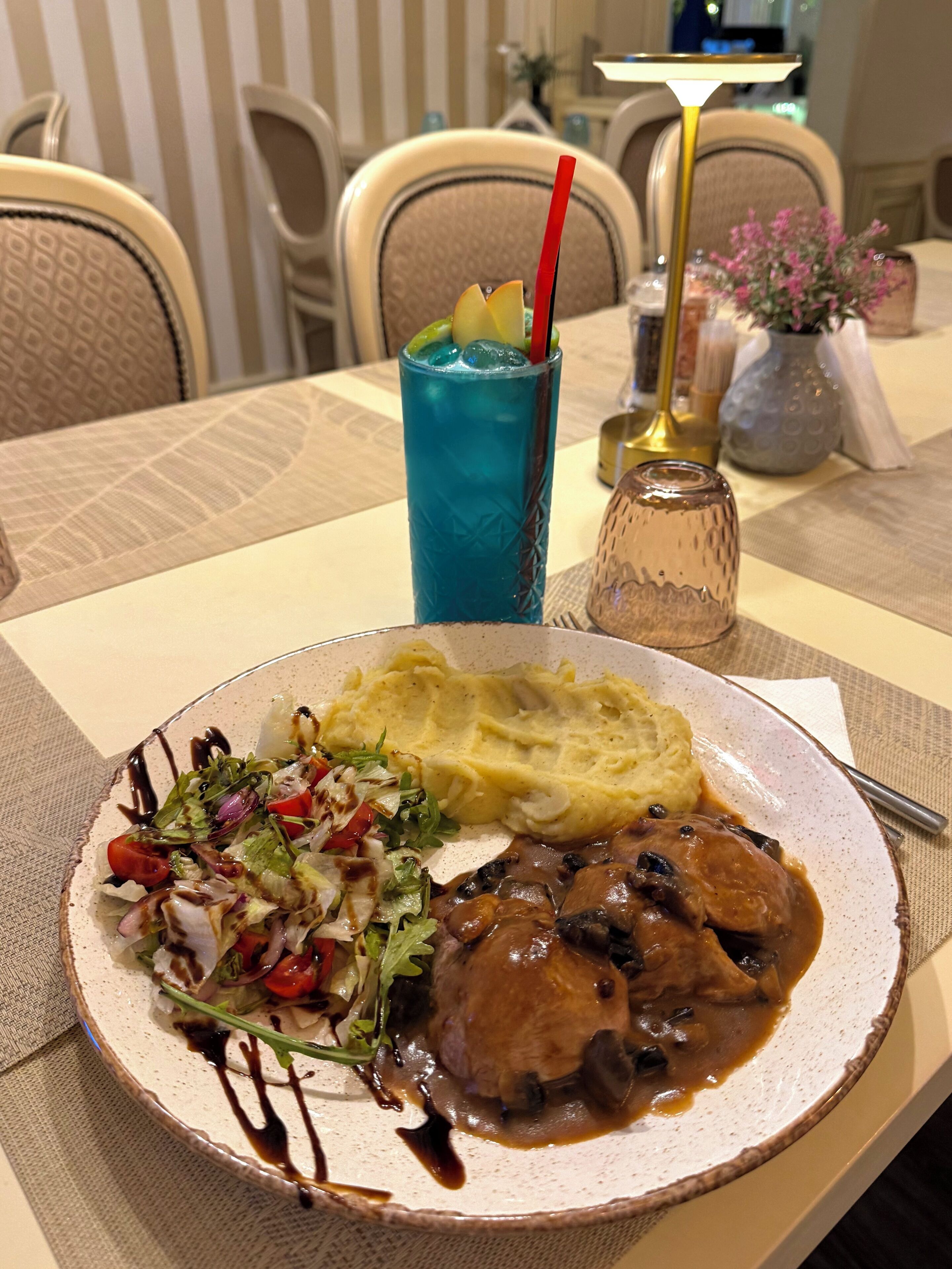 Une assiette de nourriture comprenant de la purée de pommes de terre, une salade et un plat de viande, une boisson bleue servie avec une paille, et un salier posé sur une table.