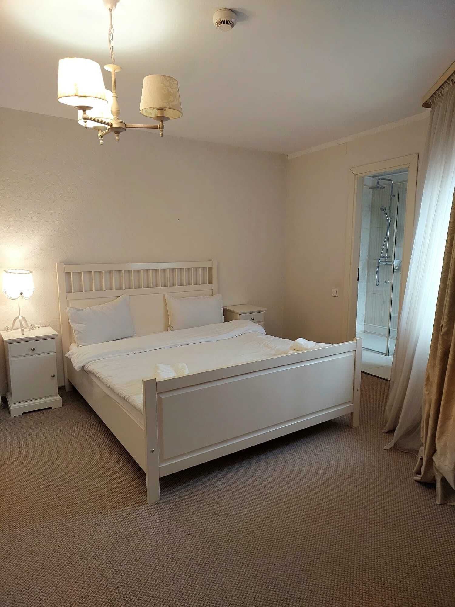 A bedroom with a bed, nightstands, a chandelier, a window with curtains, and a shower area.