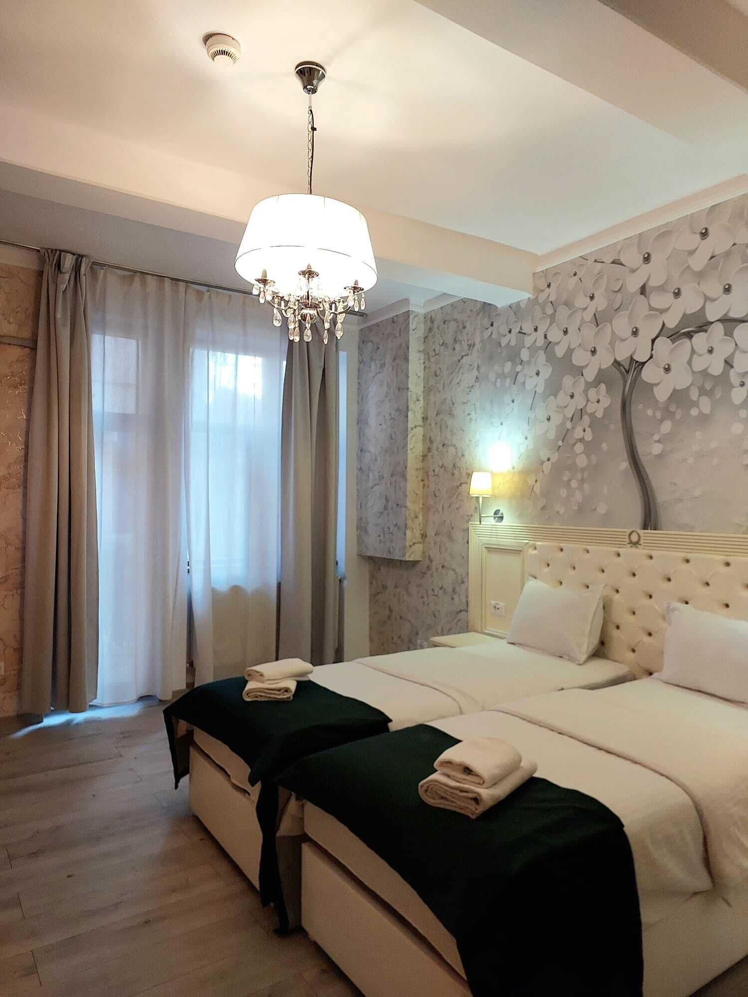 A hotel room with a large bed, two bedside tables, a chandelier, a wall with a floral mural, and a window with curtains.
