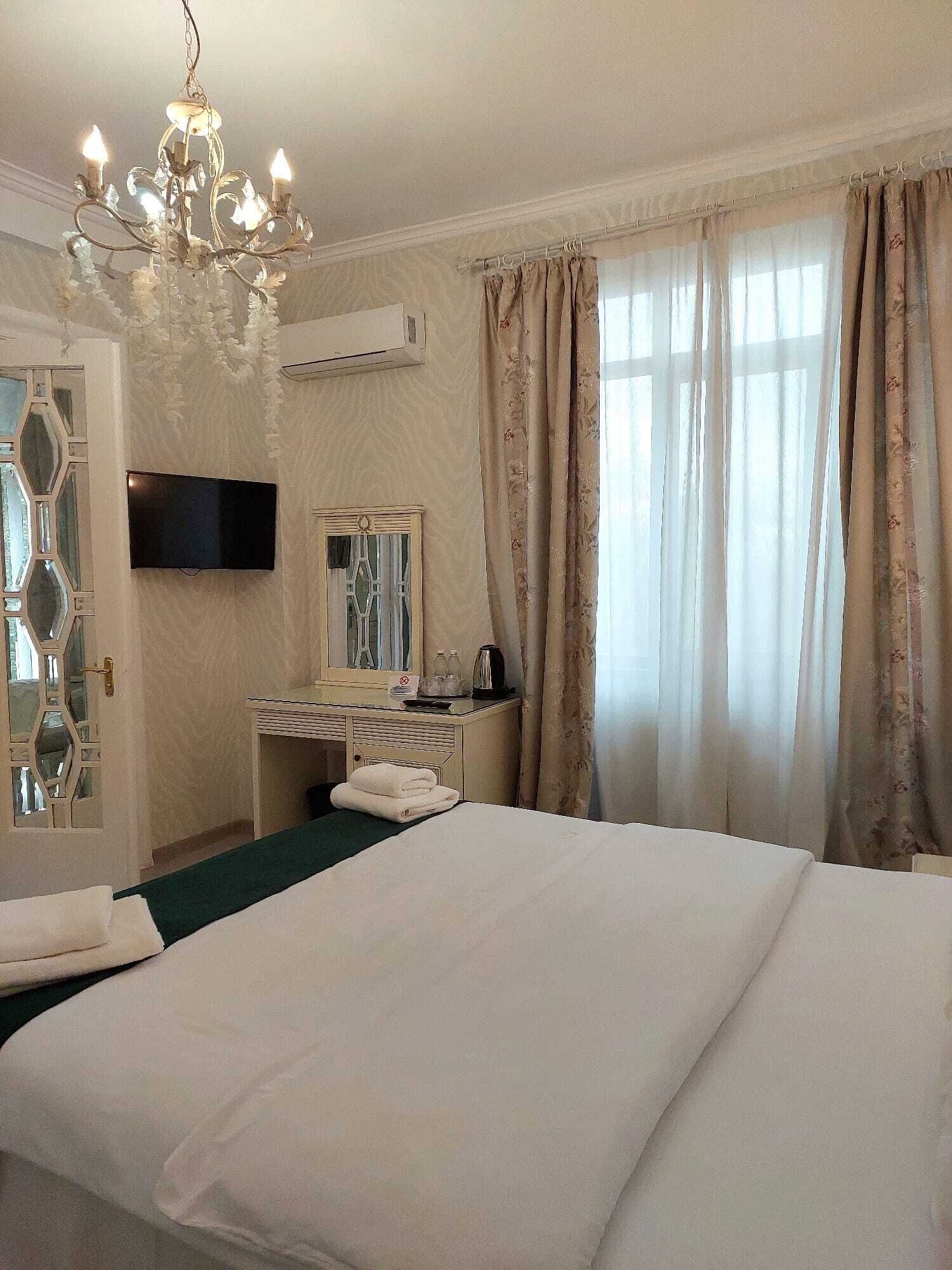 A hotel room with a large bed, a chandelier, a flat-screen TV, a desk with a mirror, and a window with curtains.