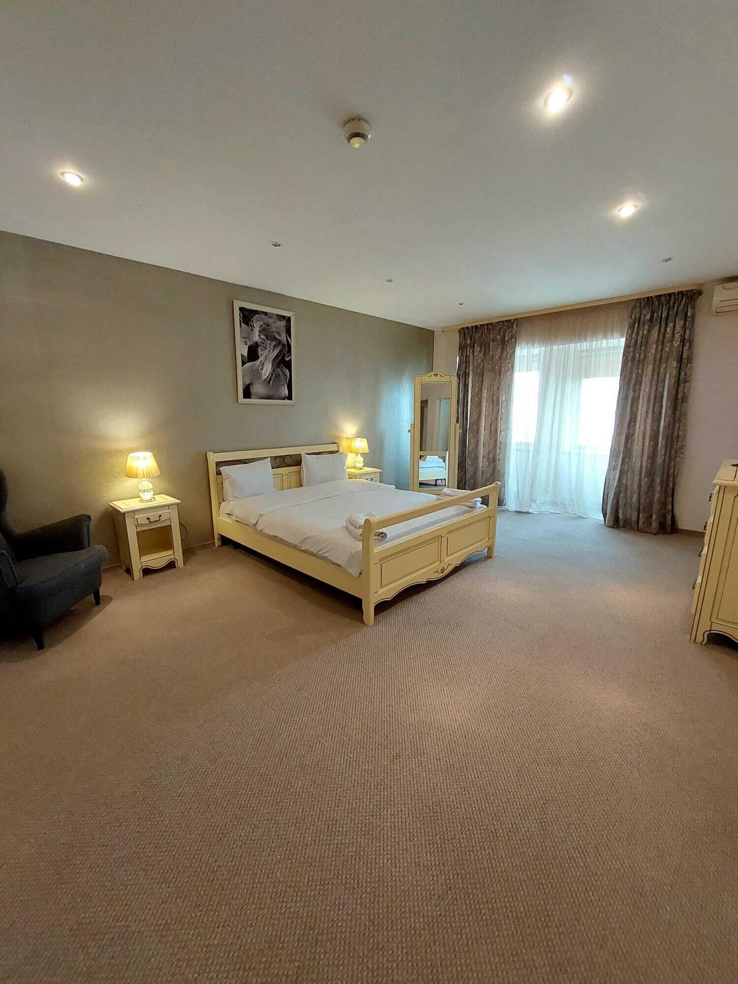 A hotel room with a bed, two bedside lamps, a chair, a mirror, and a window with curtains.