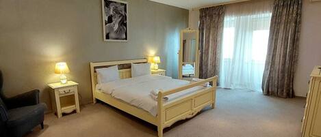 A hotel room with a bed, two bedside lamps, a chair, a mirror, and a window with curtains.