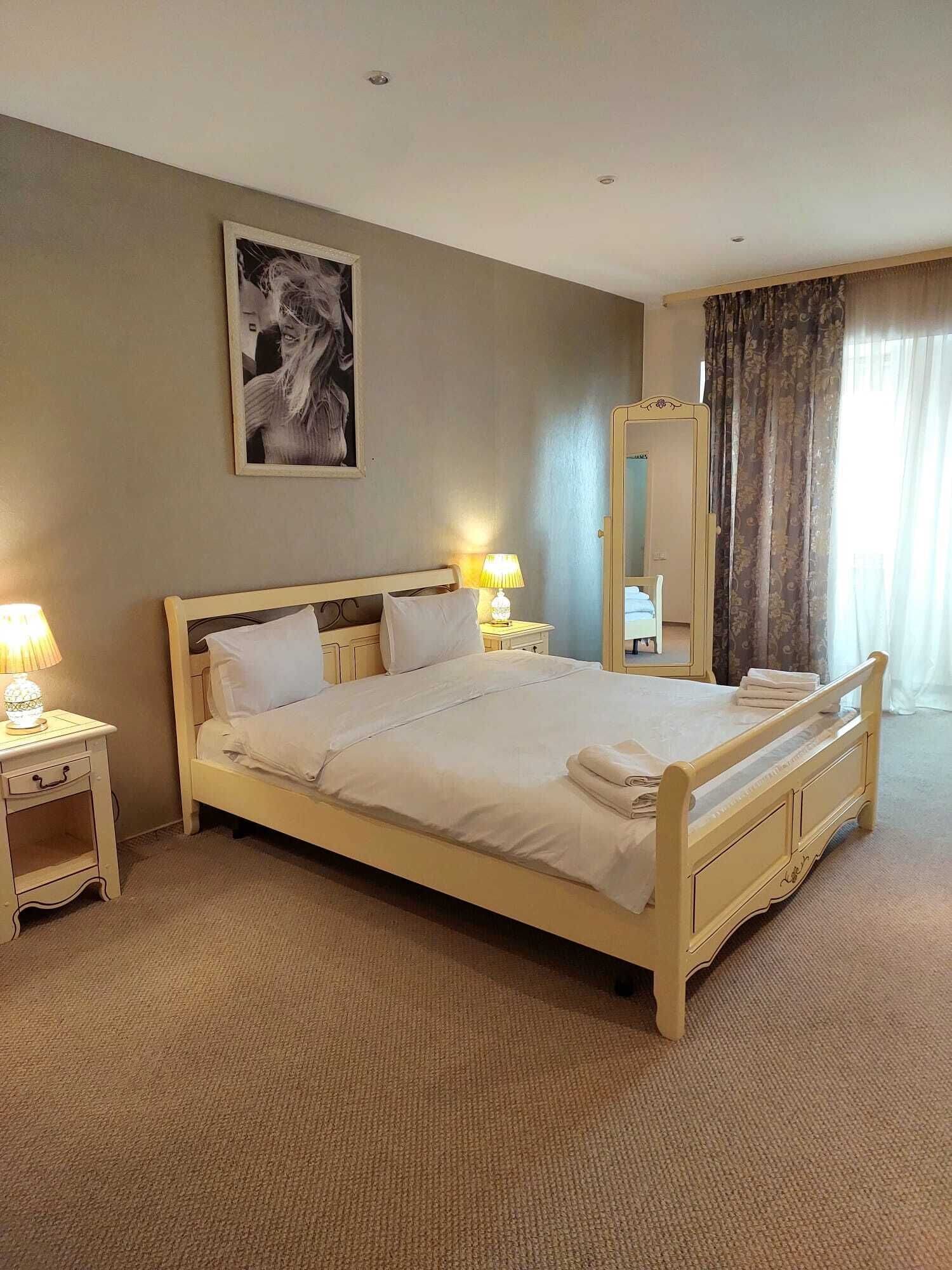 A neatly made bed with a headboard, two bedside tables with lamps, a large mirror, and a framed picture on the wall.