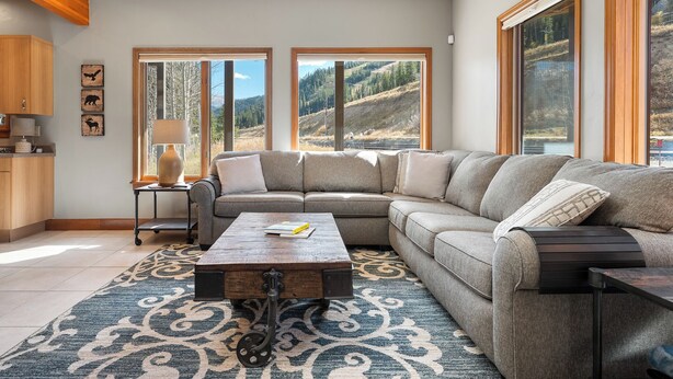 Ein modernes Wohnzimmer mit einer Eckcouch, einem Holztisch und großen Fenstern mit Blick auf eine gebirgige Landschaft.