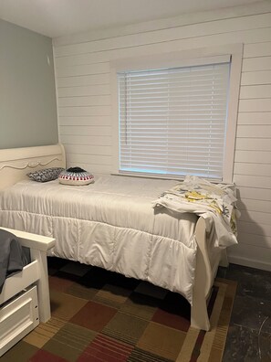 Ein Schlafzimmer mit einem Bett, einem Fenster mit Jalousien, einem gemusterten Teppich und einem weißen Kopfteil.