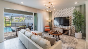 A modern living room with a large TV, comfortable seating, a dining area, and a view of a swimming pool. - Disney 5BR Villa w/ Pool Solterra Resort (Davenport)