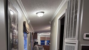 En smal hall med trägolv, en grå vägg och ett tak med en infälld lampa. I bakgrunden står en grå soffa, till höger finns en vit apparat och till vänster hänger en inramad tavla.