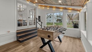 A room with a wooden-floored game table, white walls, and large windows offering a view of the outside.