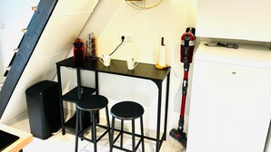 A compact kitchenette with a small table, two stools, a coffee maker, a vacuum cleaner, and a wall-mounted shelf with decorative items.