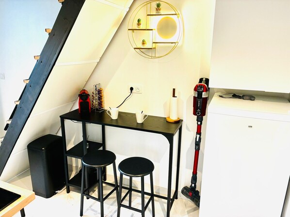 A compact kitchenette with a small table, two stools, a coffee maker, a vacuum cleaner, and a wall-mounted shelf with decorative items.