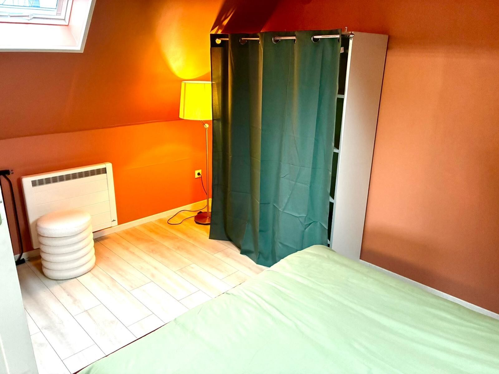 Een kleine, helder verlichte kamer met een enkel bed, een radiator, een toilet en een gordijn.