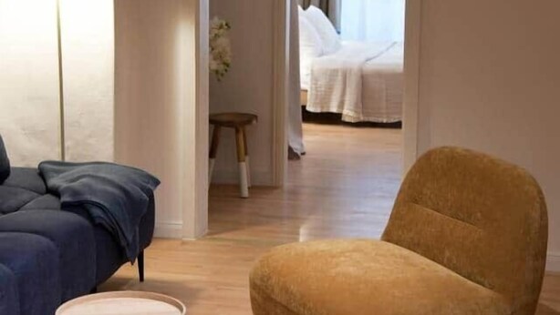 Une chambre d’hôtel moderne avec un canapé bleu, une table basse en bois et un fauteuil de couleur moutarde. Un arc en plein cintre mène à une chambre à coucher équipée d’un lit et d’une table de chevet.