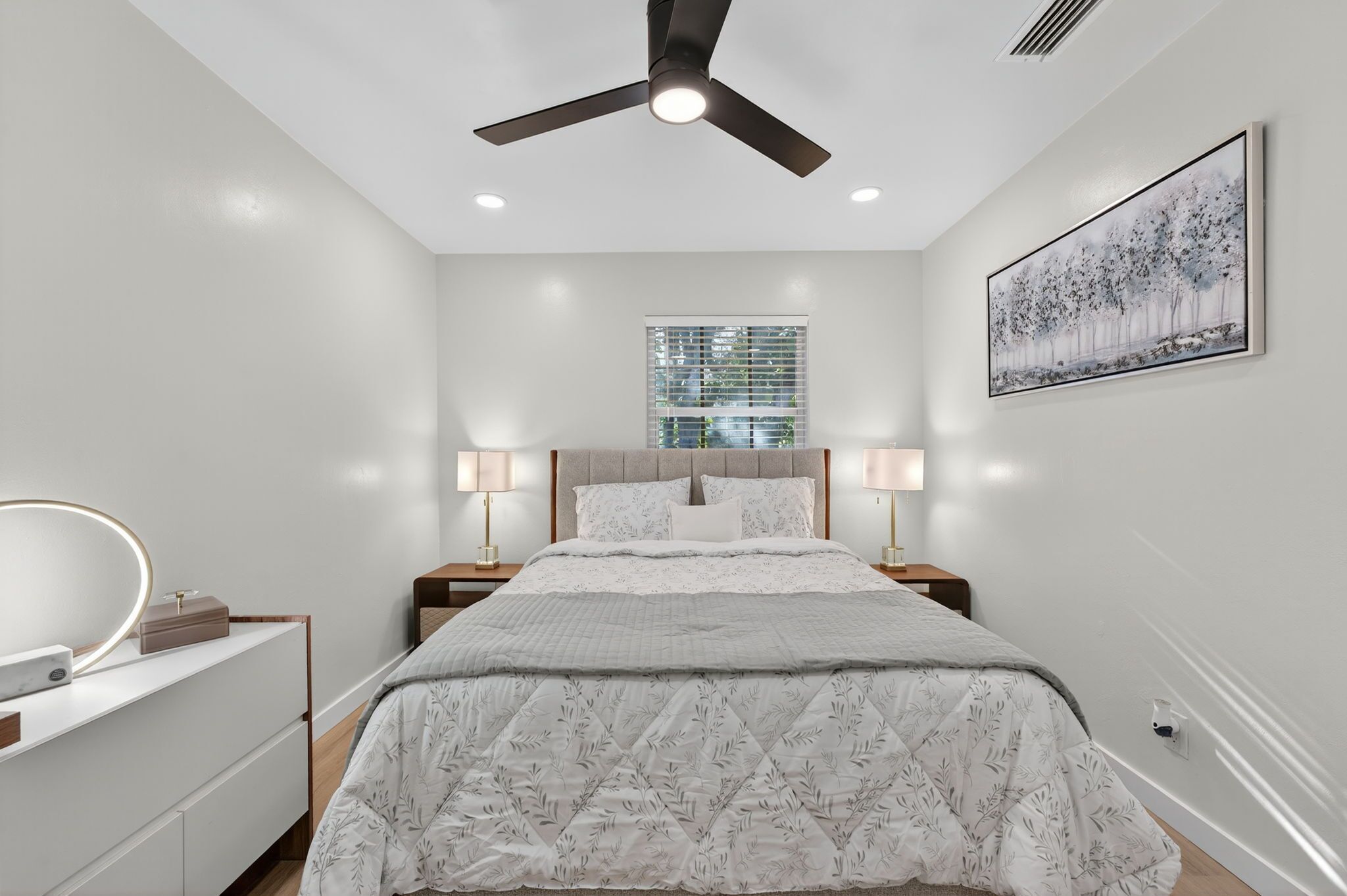 Une chambre à coucher comprenant un lit, des lampes de chevet, un ventilateur de plafond, une fenêtre et un tableau accroché au mur.