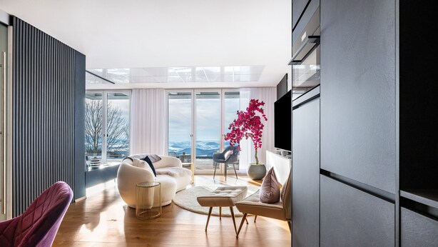 A modern living room with a sofa, ottoman, and a chair, a large window with a view, and a potted plant. - Apartment “Himmelweit” with Mountain Views, Private Terrace and Wi-Fi (Freyung)