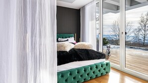 A modern bedroom with a green upholstered bed, a dark headboard, and a view of a snowy landscape through a glass door. - Apartment “Himmelweit” with Mountain Views, Private Terrace and Wi-Fi (Freyung)