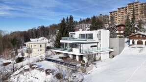 A modern two-story building with large windows and a balcony, surrounded by snow-covered trees and other buildings. - Apartment “Himmelweit” with Mountain Views, Private Terrace and Wi-Fi (Freyung)