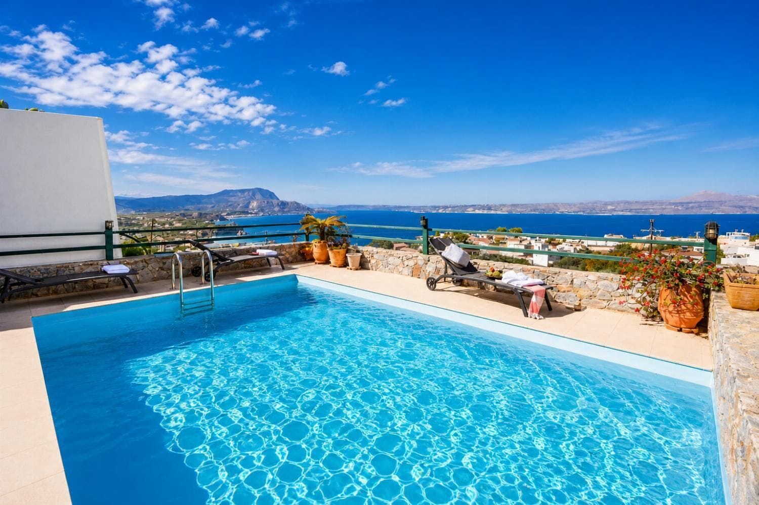 A pool with clear blue water, surrounded by lounge chairs and potted plants, overlooking a coastal landscape with buildings and a mountain in the distance.