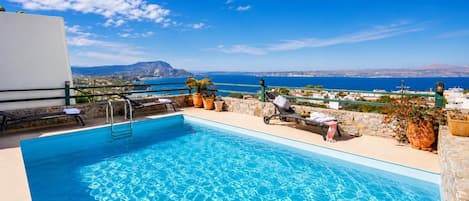 A pool with clear blue water, surrounded by lounge chairs and potted plants, overlooking a coastal landscape with buildings and a mountain in the distance.