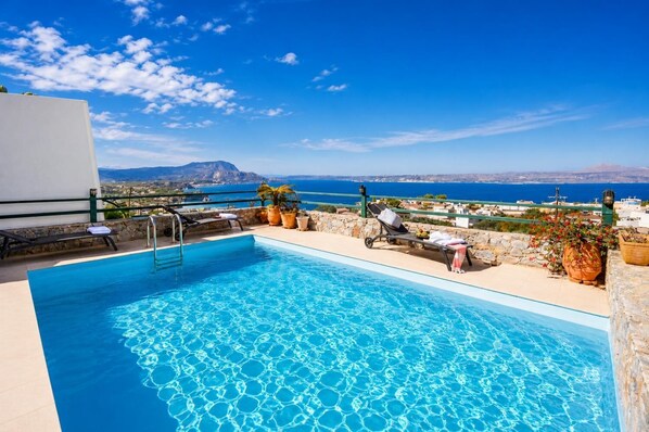 Une piscine à l’eau d’un bleu cristallin, entourée de chaises longues et de plantes en pot, d’où l’on peut admirer un paysage côtier avec des bâtiments et une montagne à l’horizon.