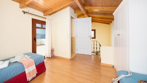 Quarto com teto de madeira, cama com colcha azul e vermelha, guarda-roupa branco e vista pela janela.