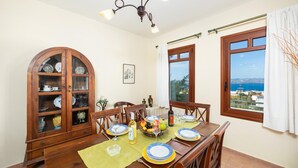 Uma área de refeições com uma mesa de madeira posta para quatro pessoas, um armário com louças e vista para o mar pela janela.