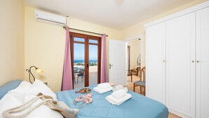 Quarto de hotel com cama, uma mesa, uma cadeira, ar condicionado e vista para o mar através de uma porta de correr.