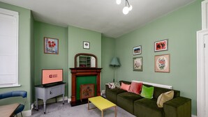 Een woonkamer met een groene bank, een gele salontafel, een open haard en ingelijste foto’s aan de wanden.