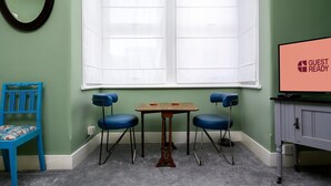 Een kamer met een groene wand, een wit raam, een houten tafel met vier blauwe stoelen, een blauwe stoel met een gestreept kussen, en een televisie waarop staat 