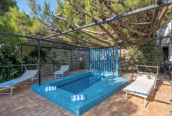 A rectangular swimming pool with blue tiles, surrounded by lounge chairs and a pergola structure.