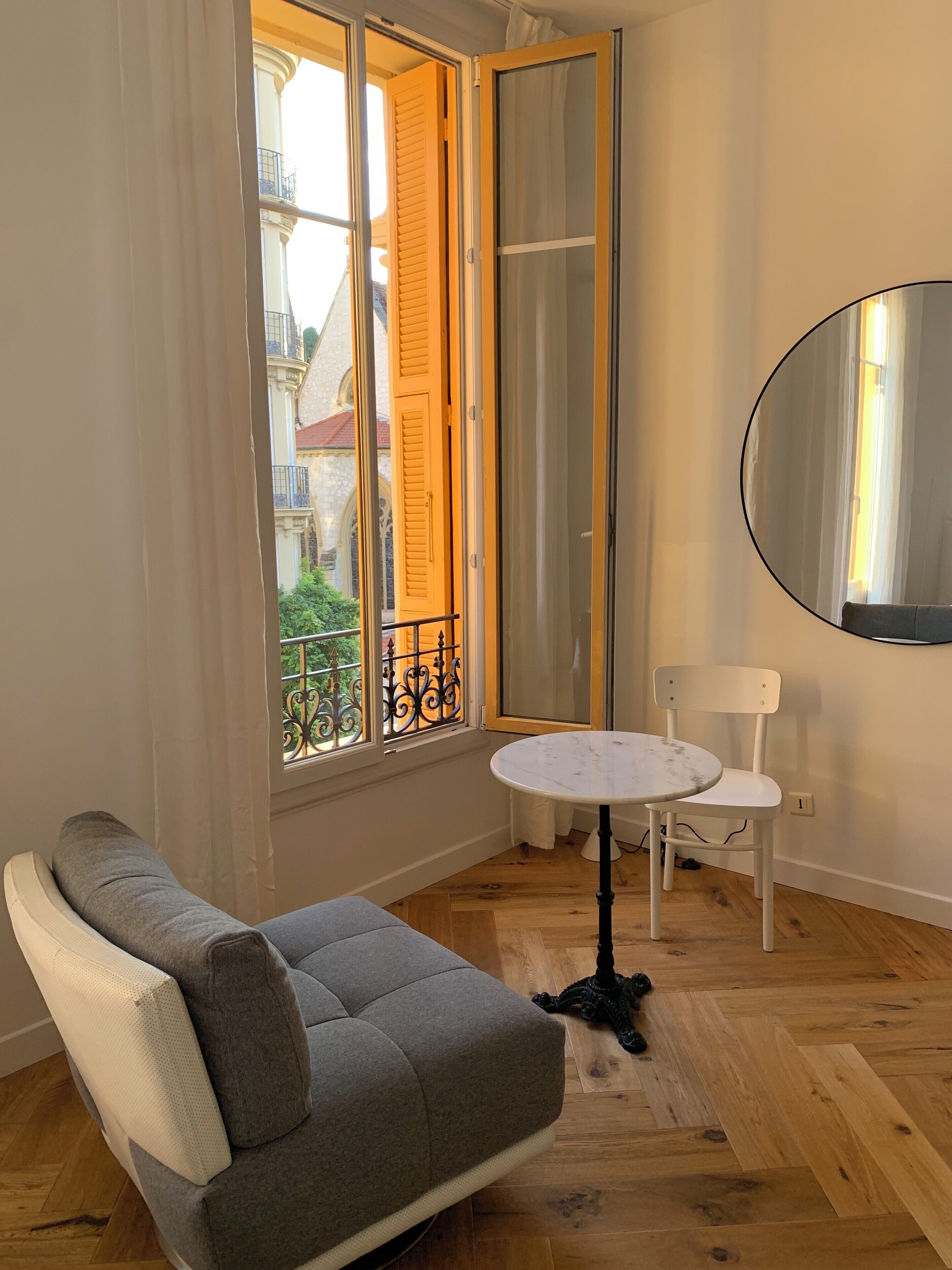 A room with a grey sofa, a small round table, a white chair, and a large mirror on the wall.