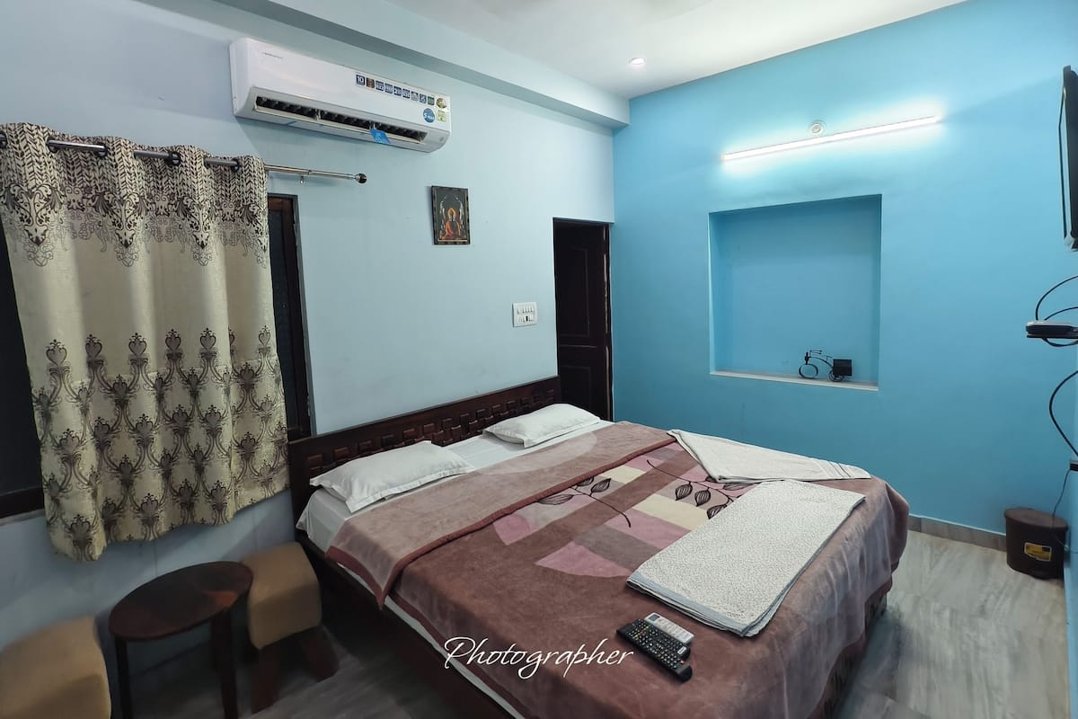 A bedroom with a bed, a bedside table, an air conditioner, a ceiling fan, a wall-mounted shelf, and a window with curtains.