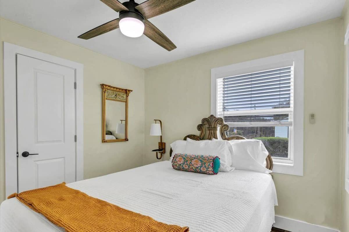 Un dormitorio con cama, ventilador de techo, ventana con persianas, puerta y espejo en la pared.