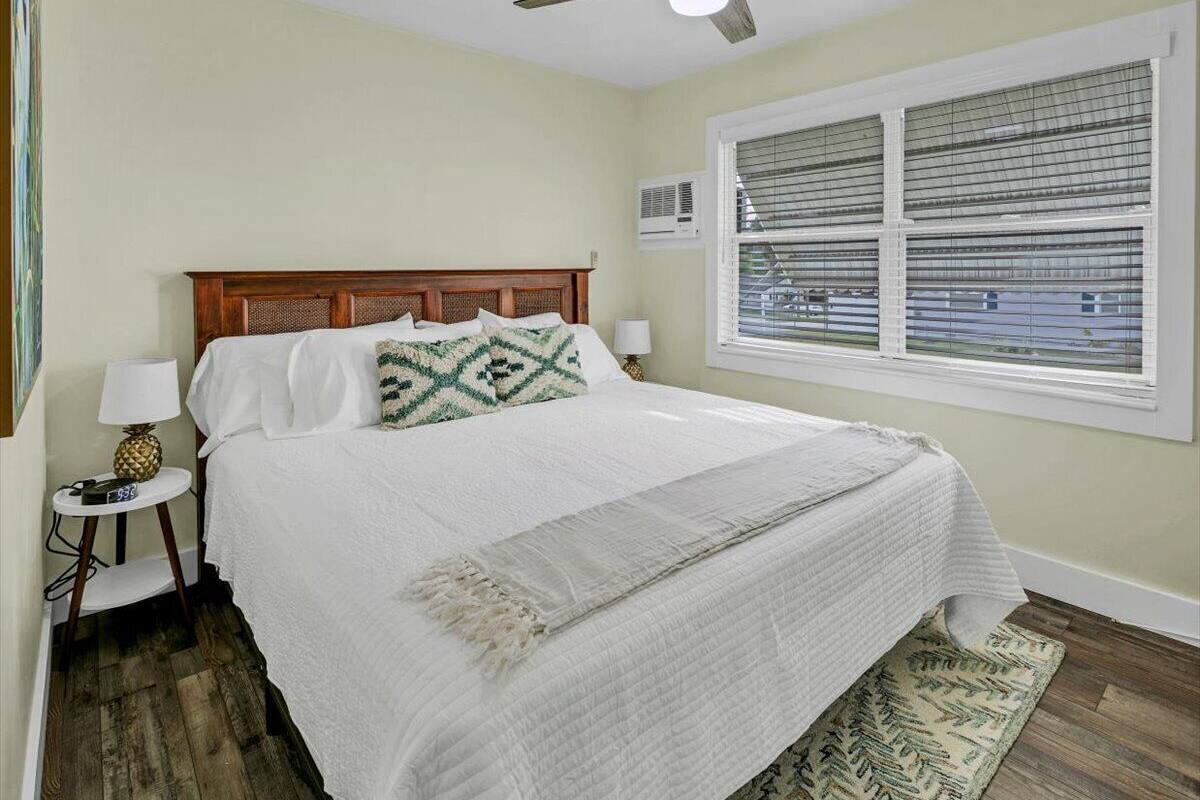 Un dormitorio con cabecera de madera, cama con ropa blanca, ventilador de techo, ventana con persianas, una mesita de noche con lámpara y una alfombra estampada.