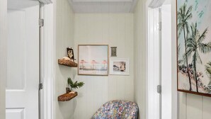 A hallway with a wooden floor, a patterned armchair, a white pouf, and wall-mounted shelves with framed pictures. - Safety Harbor Motel (Safety Harbor)