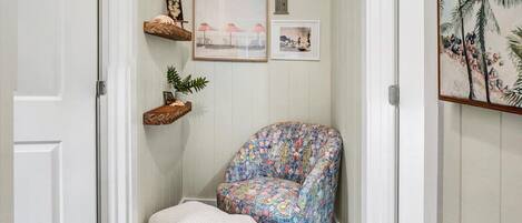 A hallway with a wooden floor, a patterned armchair, a white pouf, and wall-mounted shelves with framed pictures.