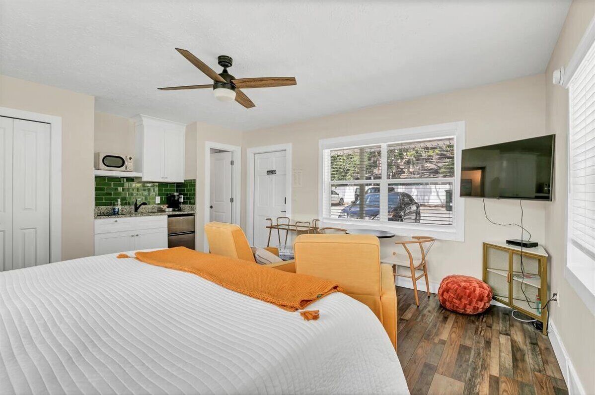 A bedroom with a bed, a ceiling fan, a TV, a window with a view of parked cars, and a kitchenette with a microwave.